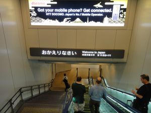 나리타 국제공항(成田国際空港) 도착 게이트의 안내판. 다른 언어는 일본 입국을 환영하는 문구인 반면, 일본어는 "어서 오세요"(おかえりなさい)라고 귀국을 환영하는 표현을 사용하고 있다.
