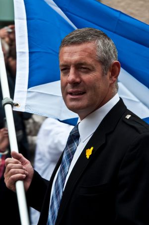 양복과 넥타이를 착용하고 스코틀랜드 국기를 들고 있는 중년 남성.