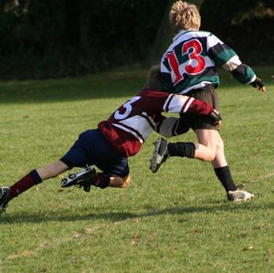 녹색과 검은색 줄무늬 럭비 저지를 입고 카메라에서 멀어지는 아이가 상대편 유니폼을 입은 다른 아이에게 엉덩이와 다리 주변을 태클 당하고 있다.