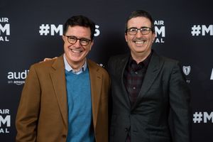 어두운 정장을 입은 올리버와 베이지색 정장을 입은 콜베어가 나란히 서서 웃고 있는 사진. 둘 다 배경 앞에 서 있다.