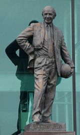 정장을 입은 대머리 남성의 청동상. 오른손은 오른쪽 허리에, 왼손은 왼쪽 허리에 축구공을 들고 있다.