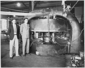 M. Stanley Livingston과 어니스트 로런스(오른쪽)가 로런스 방사선 연구소의 69 cm 사이클로트론 앞에 서 있다. 휘어진 금속틀이 자석의 코어이고, 큰 원통형 상자에는 자기장을 생성하는 와이어 코일이 들어 있다. "디" 전극이 들어 있는 진공 챔버는 자석의 극 사이 중앙에 있다.