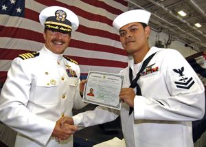 미 해군 제복을 입은 두 명의 남성이 미국 국기 앞에서 손을 잡고 시민권 증서를 들고 있다.