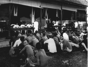 건물 앞에 쪼그리고 앉아 있는 수십 명의 남자들. 안쪽에는 흰색 옷을 입은 남자들이 책상 뒤에 앉아 있고 서 있음.