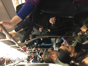 승객들로 가득 찬 Q 열차. 많은 승객들이 서로 바싹 붙어 있으며, 일부 승객은 스마트폰을 사용하고, 다른 승객들은 서서 열차 손잡이를 잡고 있다.