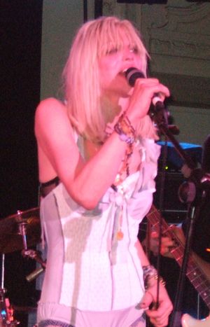 여성 뮤지션 코트니 러브가 콘서트에서 마이크에 대고 노래하고 있다. 그녀는 란제리 코르셋을 입고 있으며 긴 금발 머리를 하고 있다.