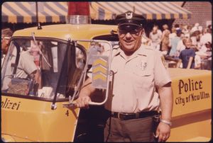 주차 미터 점검원이 독일어로 경찰을 뜻하는 단어(Polizei)가 새겨진 그의 경찰 차량 옆에 서 있습니다. 이는 독일계 민족 기원을 강조하는 마을의 일부입니다. 미네소타주 뉴울름, 1974년 7월.