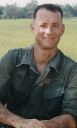 사진기 앞에서 미소를 내미는 이 사람은 파란 상자에 앉아 다리 사이로 손을 모으고 있다.