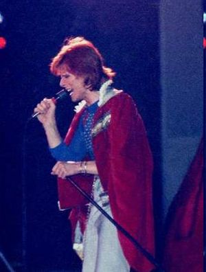 Bowie with red hair man looking to the left holding a microphone and wearing a red and white fur coat