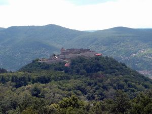 숲으로 뒤덮인 언덕 위에 자리 잡은 성, 주변은 산으로 둘러싸여 있다.