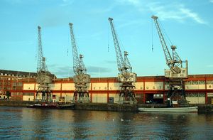 부두에 네 대의 크레인이 있는 긴 2층 건물. 두 대의 예인선이 부두에 정박해 있다.