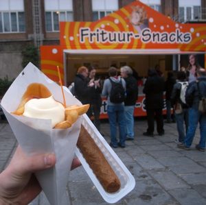 전통적인 종이 콘에 담긴 프리트(frites), 마요네즈와 커리 케첩과 함께 제공되며, 위에는 작은 플라스틱 포크가, 옆에는 프리칸델(frikandel)이 있다.