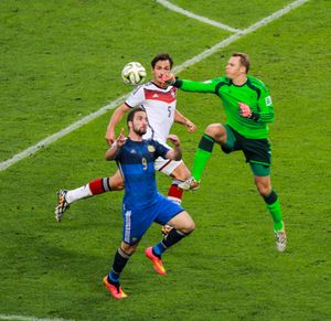 Neuer, in a Germany goalkeeper jersey, leaps into the air to punch the ball away from Argentina player Gonzalo Higuaín while Germany player Mats Hummels covers Neuer