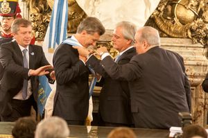 A smiling Macri puts on a blue-and-white sash with the help of Federico Pinedo and two other men