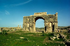 잔디밭 위에 있는 돌 아치 유적. 관광객이 아치 옆에서 포즈를 취하고 있다.