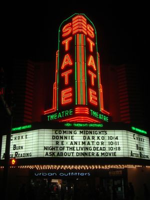 The neon marquee of a theater lists several notable cult films including Donnie Darko, Reanimator, and Night of the Living Dead.