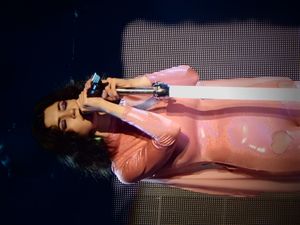 키보드를 연주하며 마이크에 대고 노래하는 젊은 갈색 머리 여성. 분홍색 유니타드를 입고 있으며, 보라색 미니 마우스 귀 모양의 머리 장식을 하고 있다.