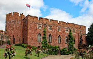 붉은 돌로 지어진 성으로 왼쪽에 포탑이 있고 접근 도로에 꽃이 늘어서 있다