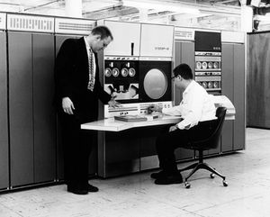 두 남자, 안경을 쓴 남자가 큰 컴퓨터 앞에 앉아있다