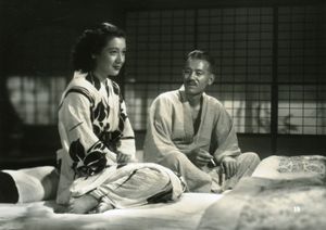 전통 의상을 입은 두 명의 일본인이 앉아 있다: 왼쪽에는 검은 머리의 젊은 여성이 오른쪽을 보고 있고, 오른쪽에는 흰머리의 노인이 여성을 보고 있다. 그들은 요에 앉아 있고 배경에는 쇼지 스크린이 있다.