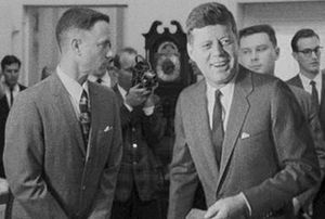 흑백 영화 스크린샷으로, 왼쪽에 주인공이 다른 남자, 케네디 대통령(배우 제드 길린의 목소리)을 바라보고 있다. 케네디는 미소를 지으며 왼쪽을 보고 있다. 배경에는 여러 남자가 각기 다른 방향을 보고 있고 한 명은 카메라를 겨누고 있다.