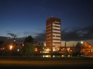 Photograph of a University campus just after sunset. A rectangular skyscraper dominates the view, standing in silhouette against a dark blue sky. To its left is a small, stone chapel with illuminated clock tower