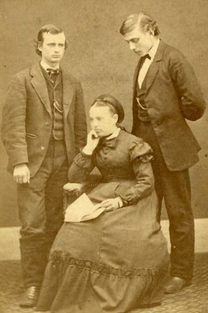 Two men standing behind a seated woman. The men are in suits and the woman is in a dress, posed with an open book on her lap.