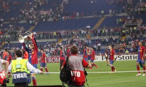 파란색과 빨간색 셔츠를 입은 남자가 머리 위로 큰 은색 트로피를 들고 있고, 다른 파란색과 빨간색 셔츠를 입은 남자들이 배경에서 지켜보고 있다.