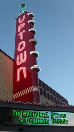 A red sign with the word "Uptown" illuminated in white neon. The sky is darkening. There is an electronic marquee that reads "Independent films screening inside."