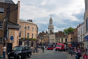 양쪽에 오래된 2~3층 건물들이 늘어선 굽어진 거리. 앞쪽에는 광고가 붙은 검은색 런던 택시가 있고, 거리 중간에는 교통량이 많은 교차로가 있다. 뒤쪽에는 돔형 시계탑이 솟아 있다.