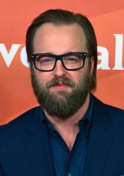 안경을 쓰고 짙은 수염과 빗질한 머리를 한 중년 백인 남자가 웃고 있으며, 짙은 파란색 정장과 셔츠를 입고 있는 사진입니다.