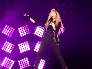 보라색 조명 앞에서 검은 옷을 입은 금발 여성이 마이크를 보며 노래를 부르고 있다.