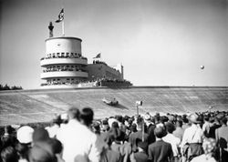 A large crowd of people watch a streamlined single-seater drive around a massive curved racing bank. A Nazi flag is flying in the distance atop a tower.