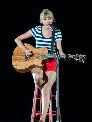 하얀색과 검은색 줄무늬 셔츠와 빨간 반바지를 입고 어쿠스틱 기타를 연주하는 스위프트