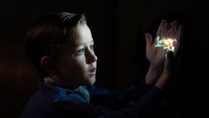 In darkness, a boy gazes intently at his palms onto which light is projected