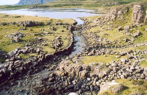 A stone lined ditch of primitive construction leads from a small lake. Rocky heathland lies on either side and there are tall cliffs in the distance.