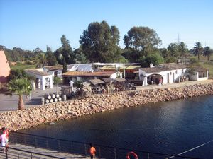 Colour photo showing the reconstruction of the ancient port of <i>Palos de la frontera</i>: transportation barrels, low buildings bordered by a covered gallery, a chariot at the very edge of the water awaiting the arrival of a boat.