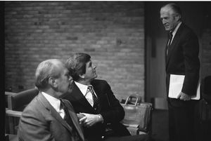 세 명의 남자가 정장을 입고 있으며, 두 명은 앉아 있고 한 명은 서 있습니다. 오른쪽에 서 있는 사람은 키가 크고 날씬하며 회색 머리입니다.