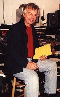 A gray-haired man with a red shirt and jacket sitting on a chair