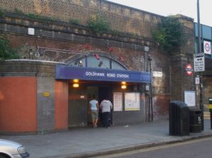 철도 벽돌 고가도로 아래 입구로, 흰색 글씨로 "GOLDHAWK ROAD STATION"이라고 적힌 파란색 표지판과 그 앞에 두 여성이 회색 하늘 아래 걷고 있다