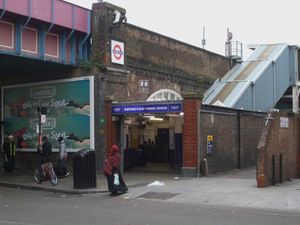 강철 다리 위에 도로를 건너는 벽돌 고가도로 위의 철도로, 흰색 글씨로 "SHEPHERD'S BUSH MARKET STATION"이라고 적힌 파란색 표지판 아래 입구가 있다