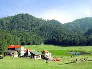 Daytime sunlit view of a verdant bowl-shaped meadow with a small pond in a valley rimmed by several prominent hills covered in evergreen forest. At centre-left and looking out on the meadow, a small cluster of tourist lodges with pitched roofs, one a bright red. Around them and the pond are small, gaily coloured gazebos and shade-giving umbrellas. Several dozen tourists and sheep mill about the pond and in the meadow.