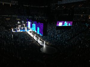 A darkened stage seen from above. Vertical and horizontal white poles of lights rise from both the main stage and from a long catwalk in front of it. Above a large screen above the catwalk shows white-bluish skyscraper images against a purplish night sky