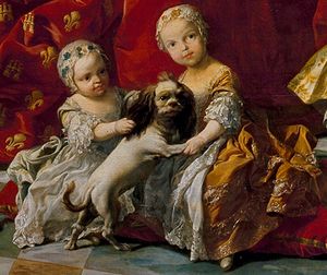 빨간색 배경 앞에서 흰색 옷을 입은 유아와 흰색과 금색 옷을 입은 유아가 작은 개와 놀고 있다