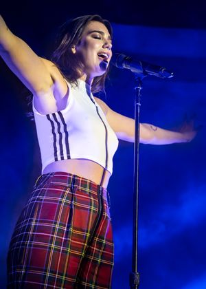 Dua Lipa with her arms out singing into a microphone. She wears a white shirt and red plaid pants.