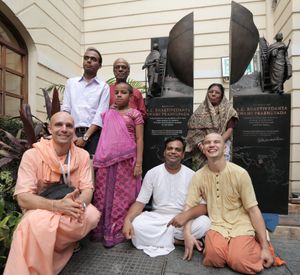 프라부파다의 아들 브린다반 찬드라 데(뒷모습, 빨간 셔츠 착용)와 그의 가족(서 있음) 및 기념비 팀(왼쪽에서 오른쪽으로 앉음): 컨셉 디렉터, 매니저 및 조각가 앞에 서 있는 Jaladuta 기념비. ISKCON 콜카타. (2015년 8월)