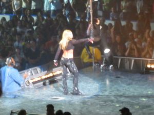 금발의 여성 공연자의 이미지. 그녀는 무대 끝 부분에서 카메라를 등지고 있다. 검은색 바지와 긴 소매가 달린 검은색 상의를 입고 허리를 드러내고 있다. 그녀 앞에는 여성 댄서가 기둥을 오르고 있다. 박수치고, 소리 지르고, 사진을 찍는 군중이 관중석에 있다.