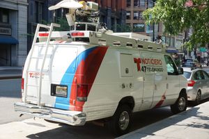 지붕에 방송 장비를 갖춘 흰색 패널 밴. 차량은 파란색과 빨간색 액센트 디자인으로 랩핑되어 있으며 측면에 Noticiero Telemundo 47 로고가 새겨져 있습니다.