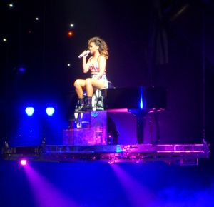 리한나가 보라색 조명으로 빛나는 무대에서 그랜드 피아노 위에 서 있다.