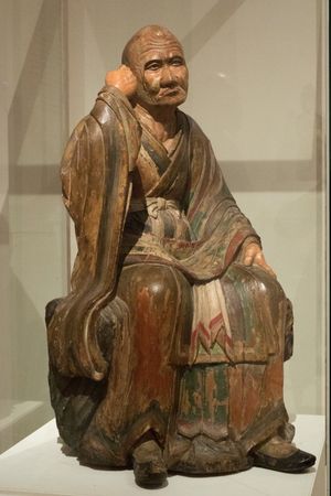 엄숙한 표정에 두개골에 약간 돌출부가 있는 승려가 앉아 지팡이를 잡는 듯 손을 들고 있다